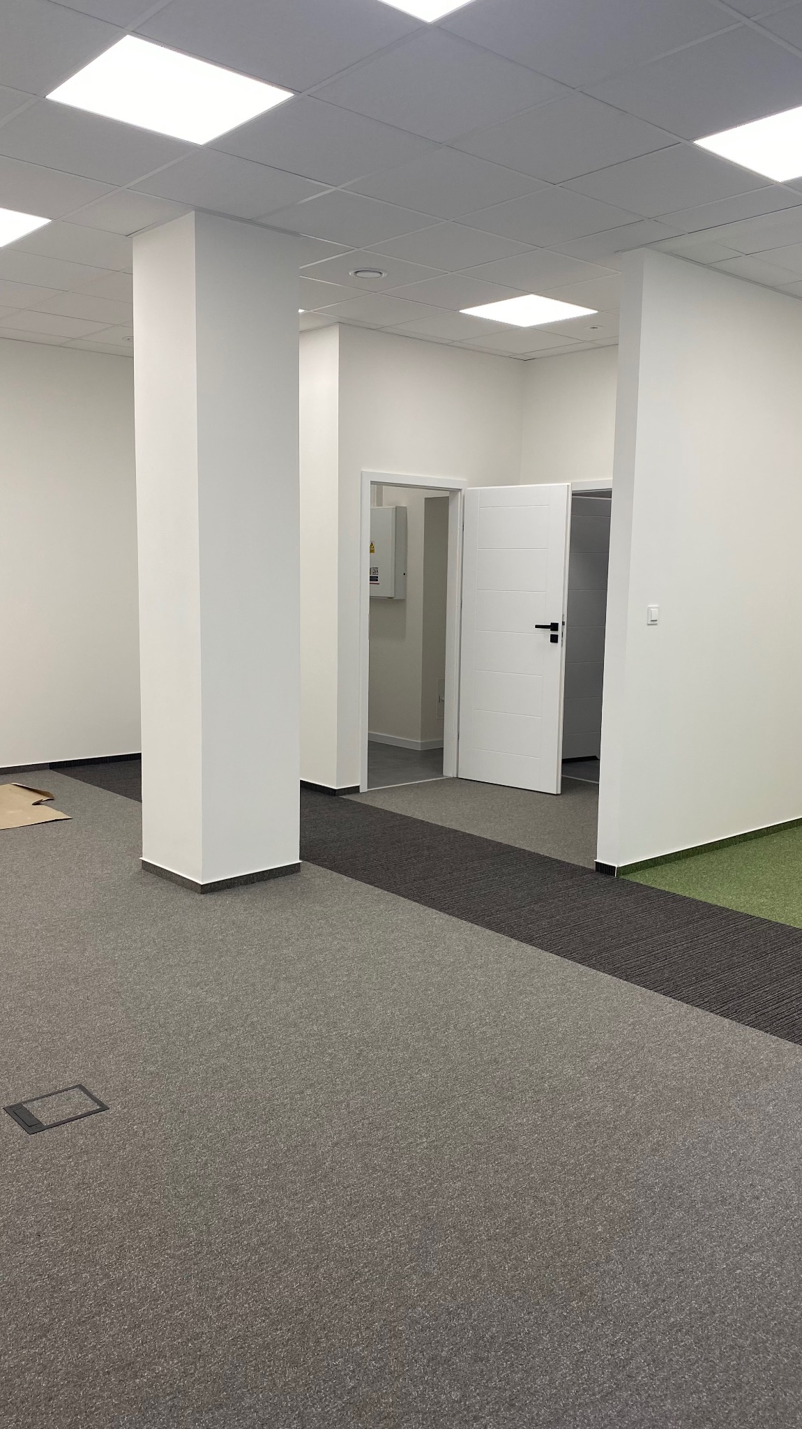 Wnętrze biurowe po remoncie: białe ściany, kolumny, sufit podwieszany z rastrowymi lampami, szara wykładzina dywanowa z ciemniejszym pasem, otwarte białe drzwi do kolejnych pomieszczeń.