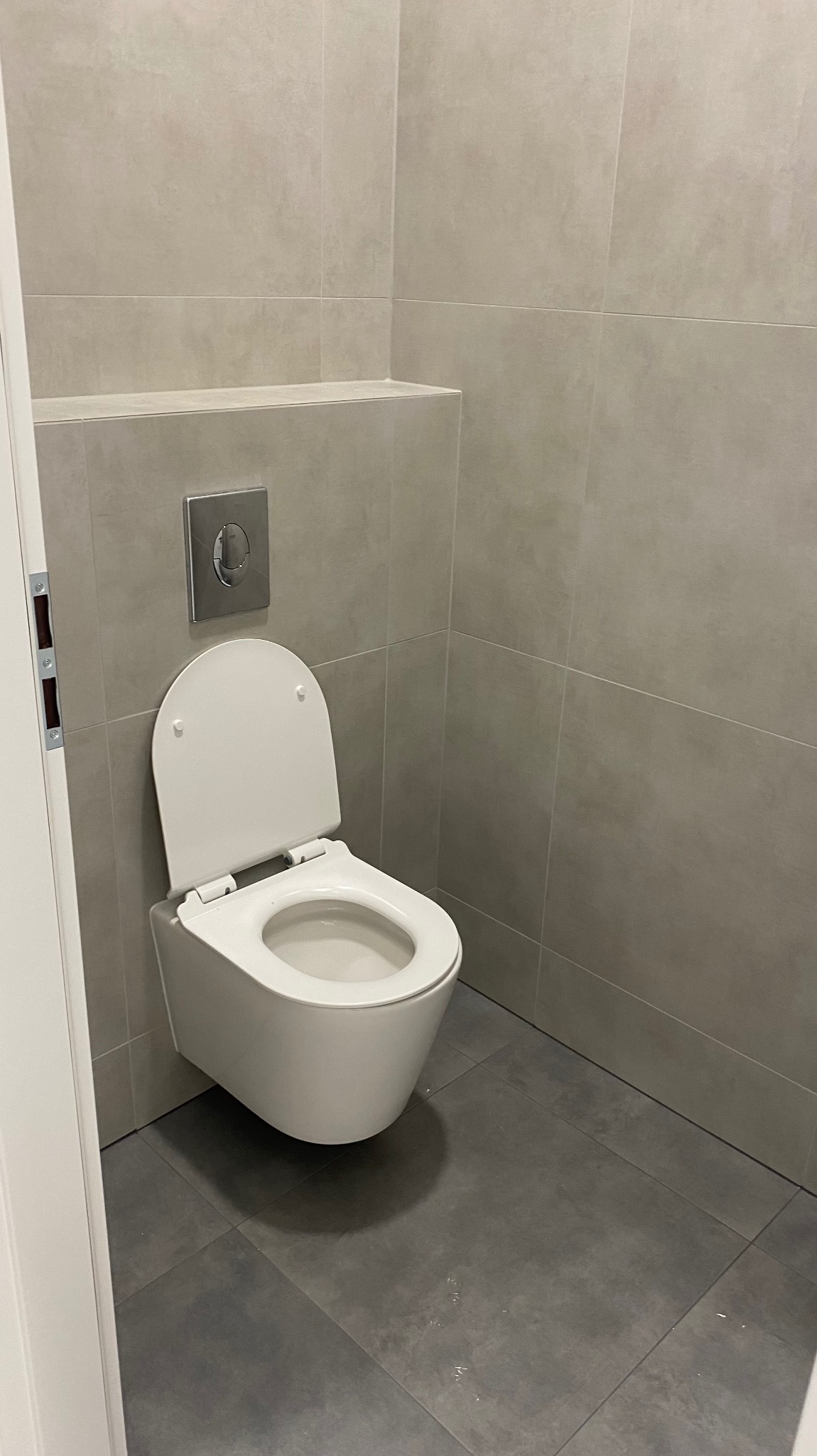 Wisząca toaleta z otwartą deską w minimalistycznej łazience wykończonej płytkami ceramicznymi w odcieniach szarości, z widocznym przyciskiem spłukującym.