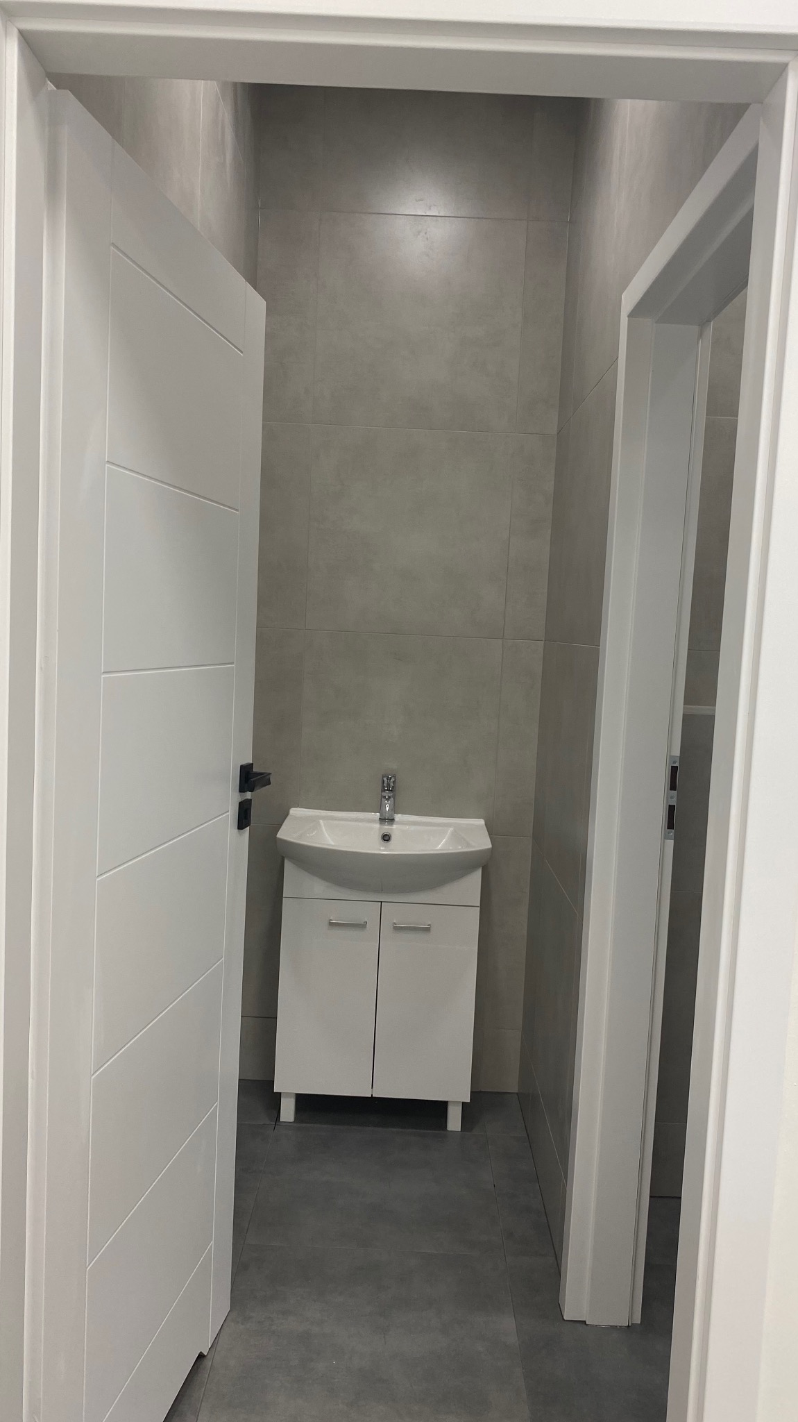 Wykończona łazienka w Warszawie z szarą płytką na ścianach i podłodze, biała szafka pod umywalką z chromowaną armaturą, widok przez otwarte drzwi.