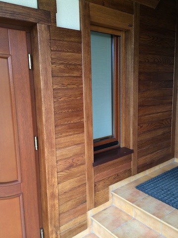 Wejście do domu z drzwiami i oknem w drewnianej ramie, obite deskami z widoczną strukturą drewna, schody wyłożone płytkami.