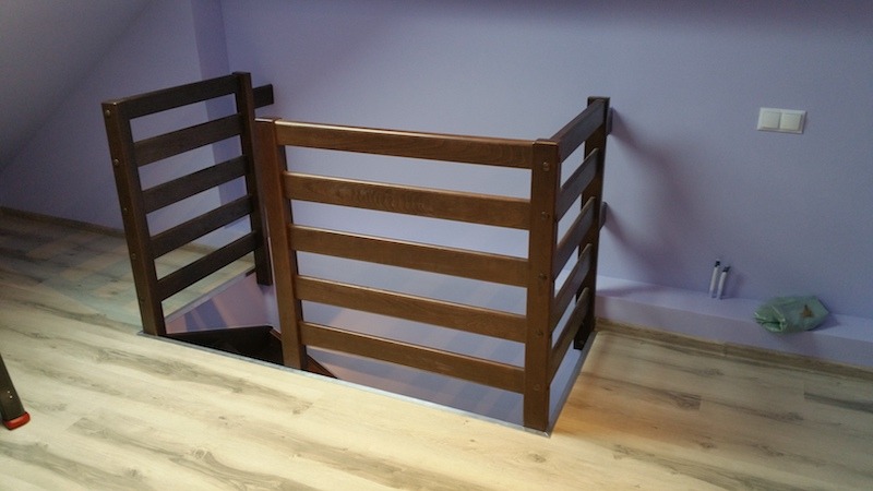 Drewniana barierka zabezpieczająca otwór schodowy na poddaszu, z poziomymi szczeblami i widocznymi śrubami, na tle fioletowej ściany i laminowanej podłogi.