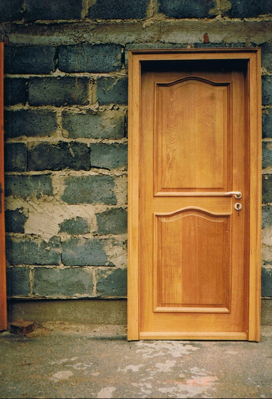 Jasne, dwupanelowe drzwi z klamką na tle surowej ściany z szarych pustaków.