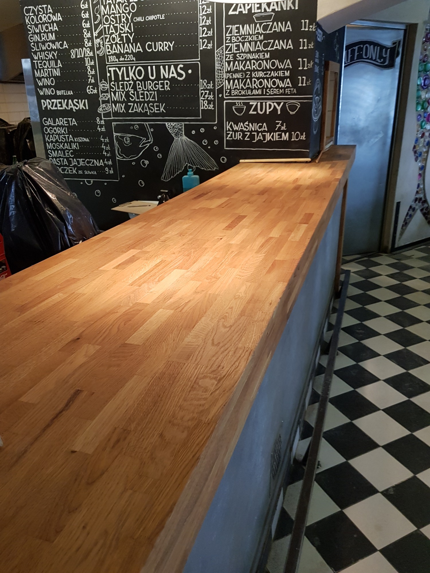 Blat baru z drewna dębowego o ciepłym odcieniu, widoczny na tle tablicy z menu i podłogi w szachownicę.