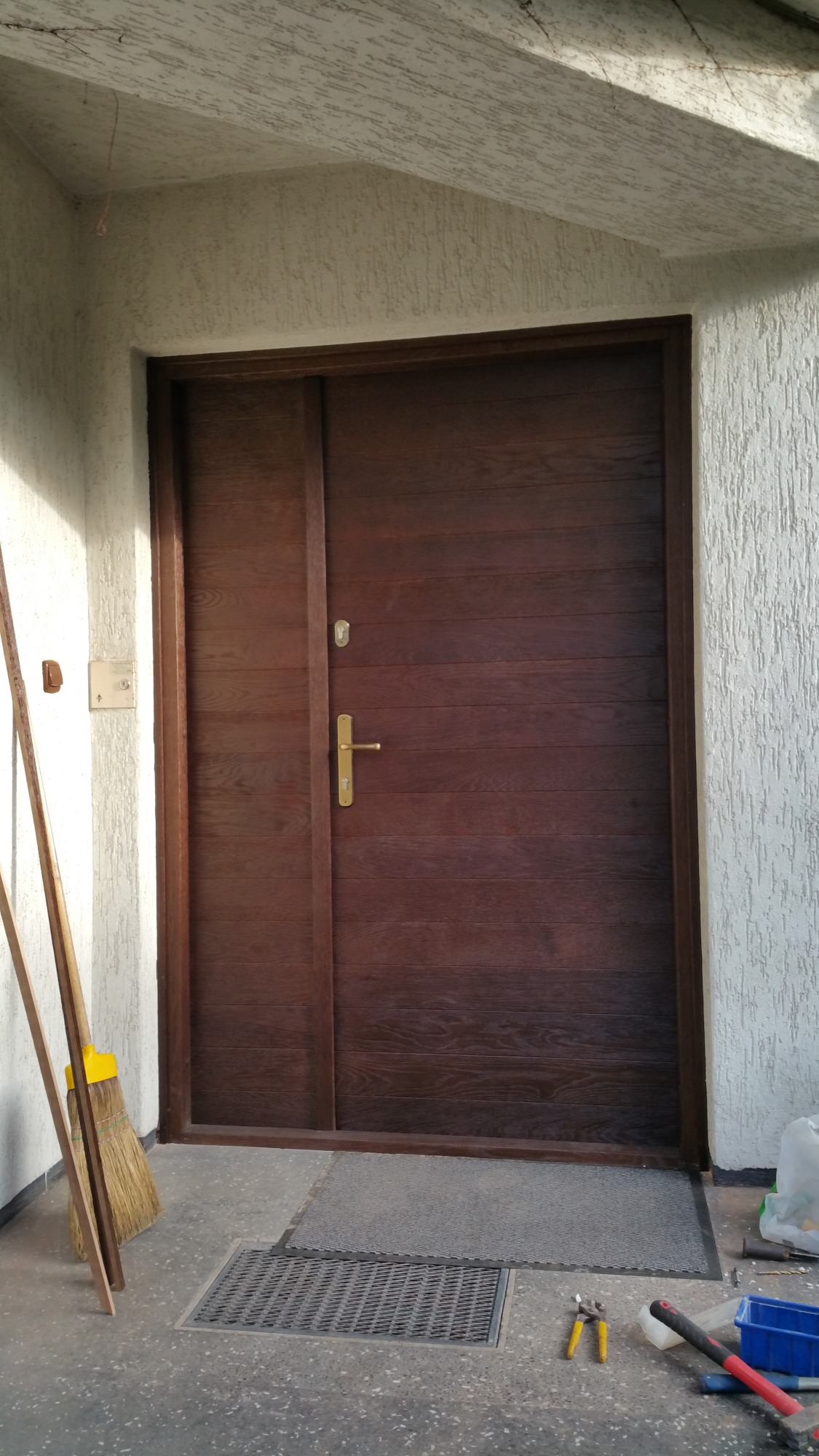 Ciemnobrązowe drzwi wejściowe z poziomym usłojeniem, złoty zamek i klamka, widoczne narzędzia i akcesoria montażowe na posadzce.