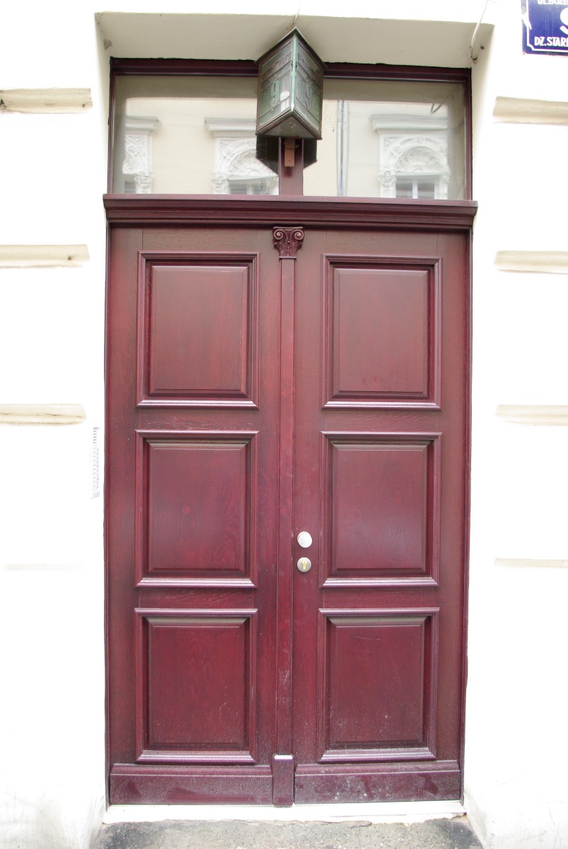 Dwuskrzydłowe, ciemnoczerwone drewniane drzwi wejściowe z prostokątnymi panelami, zwieńczone dekoracyjnym elementem architektonicznym i skrzynką na listy.