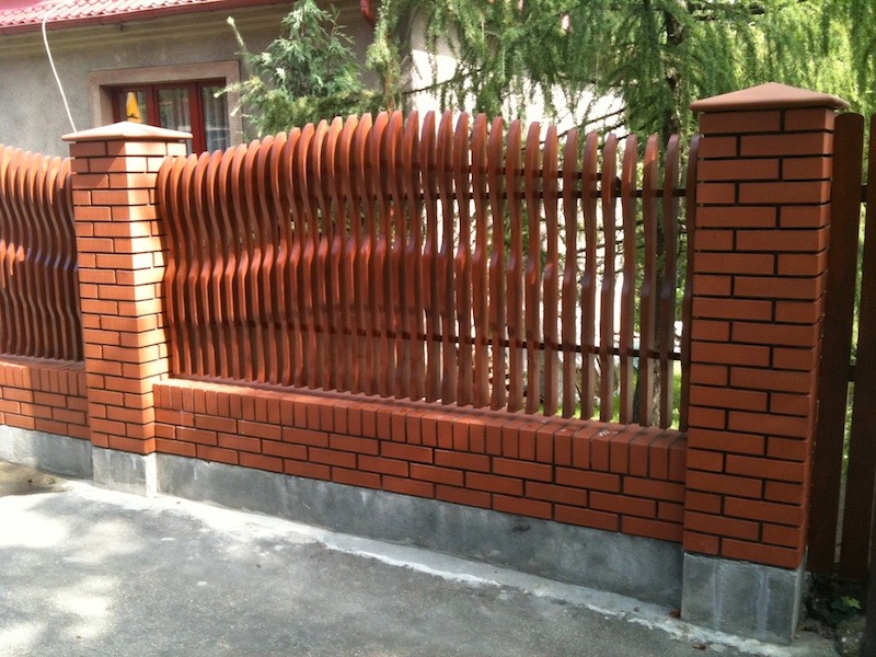 Ceglana podmurówka z oryginalnym, falistym wypełnieniem z lakierowanych listew, tworzących ogrodzenie posesji.