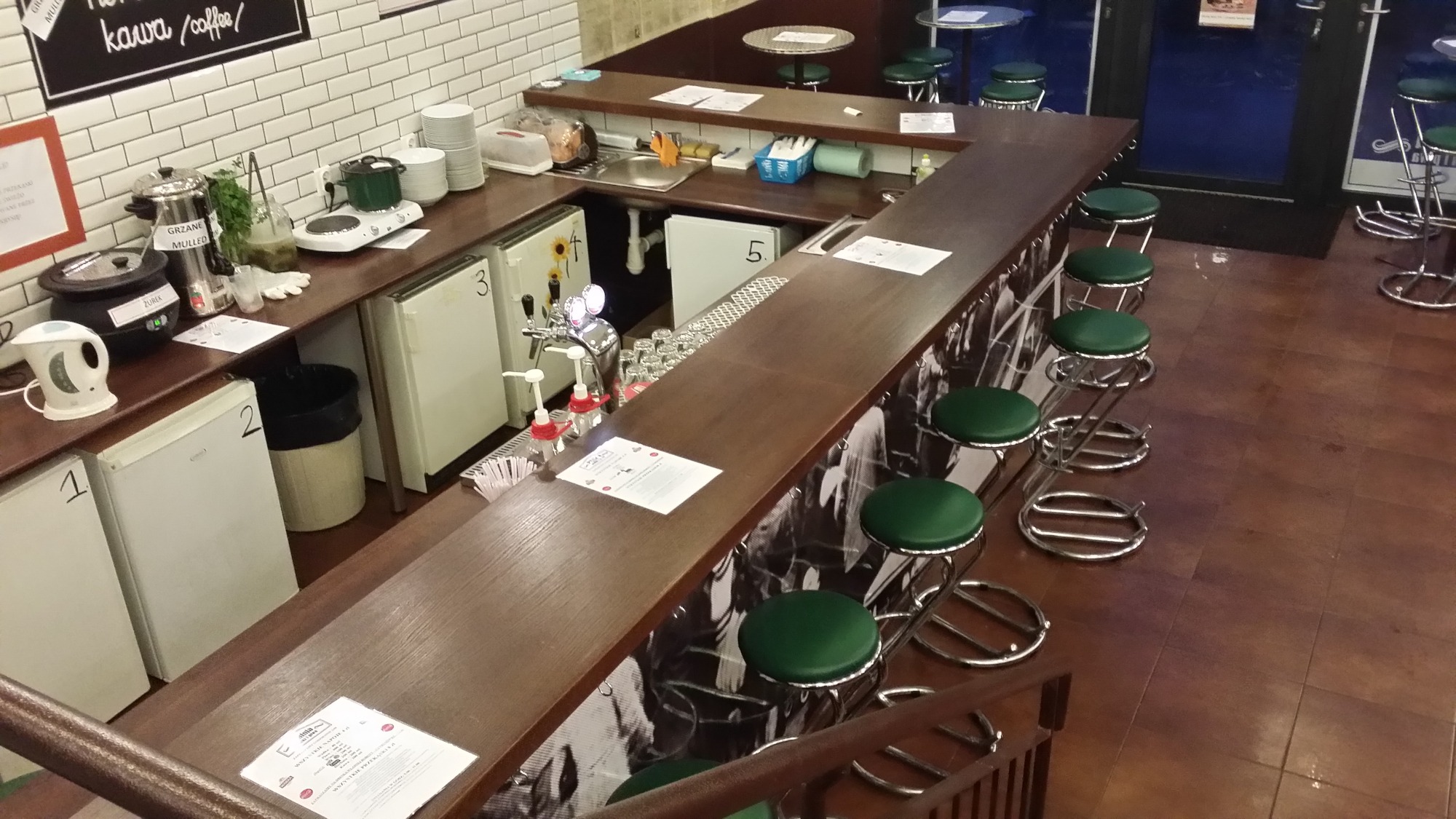 Widok z góry na długi, drewniany bar z zielonymi stołkami, wyposażony w kran do piwa, szklanki i lodówki z numerami, z widocznymi elementami zaplecza gastronomicznego, takimi jak czajnik, garnki...