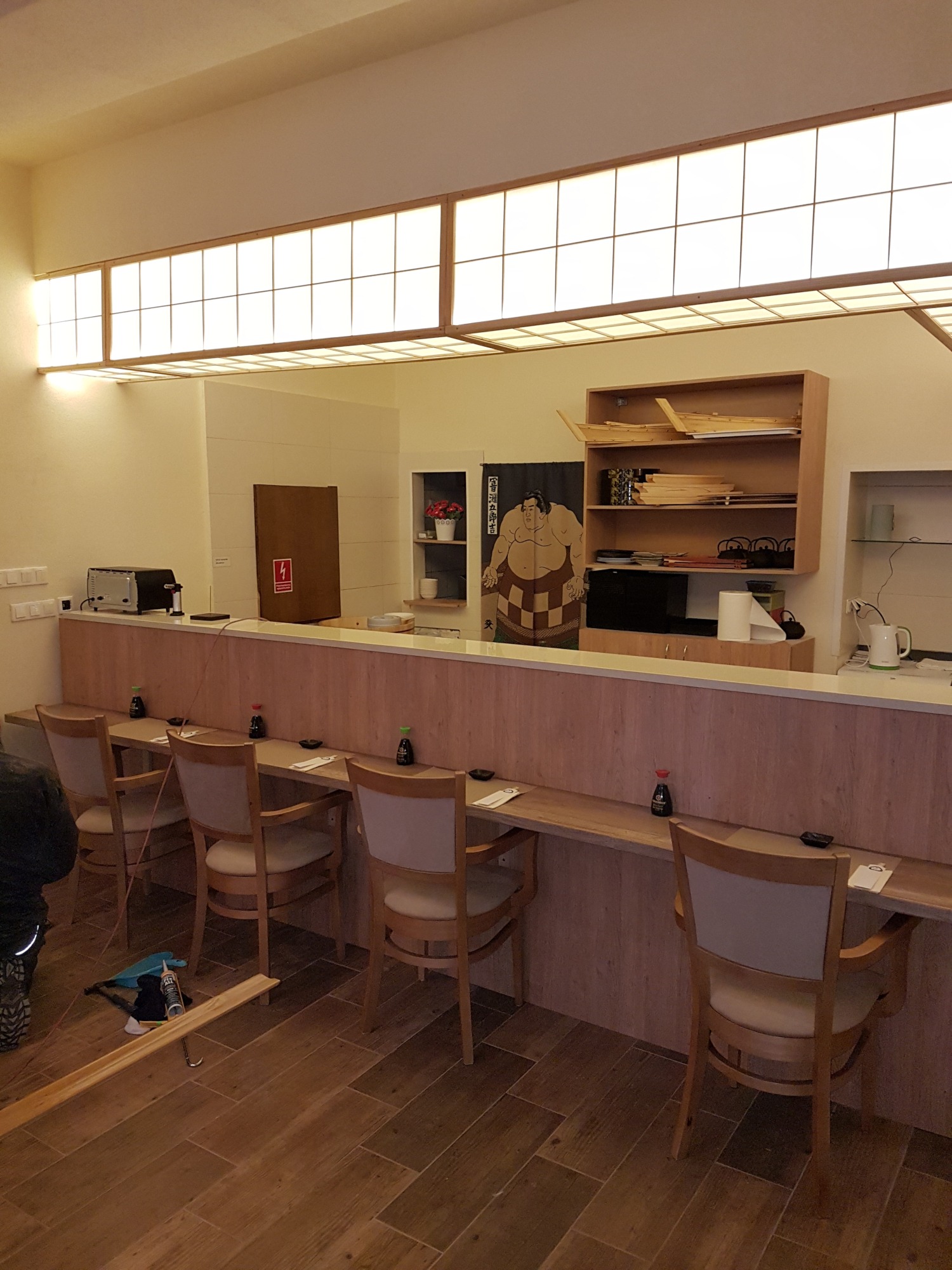 Wnętrze lokalu gastronomicznego z drewnianą zabudową: bar, krzesła z drewnianymi elementami, oświetlenie sufitowe w stylu japońskim, dekoracja z motywem sumo, narzędzia i materiały budowlane...