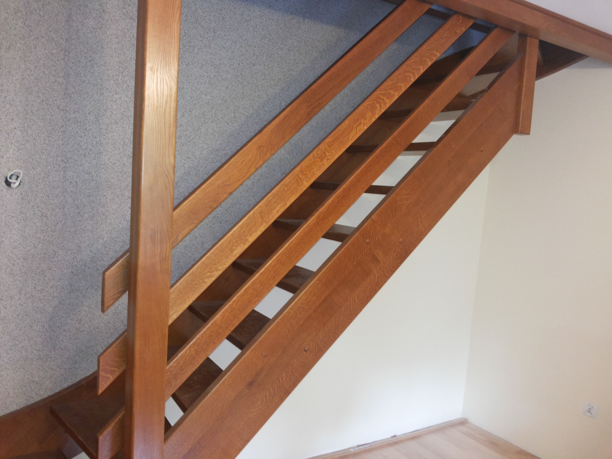 Klasyczne, brązowe schody z drewnianą balustradą z prostymi szczeblami, widok z dołu, szara ściana z widocznym gniazdkiem elektrycznym w tle.