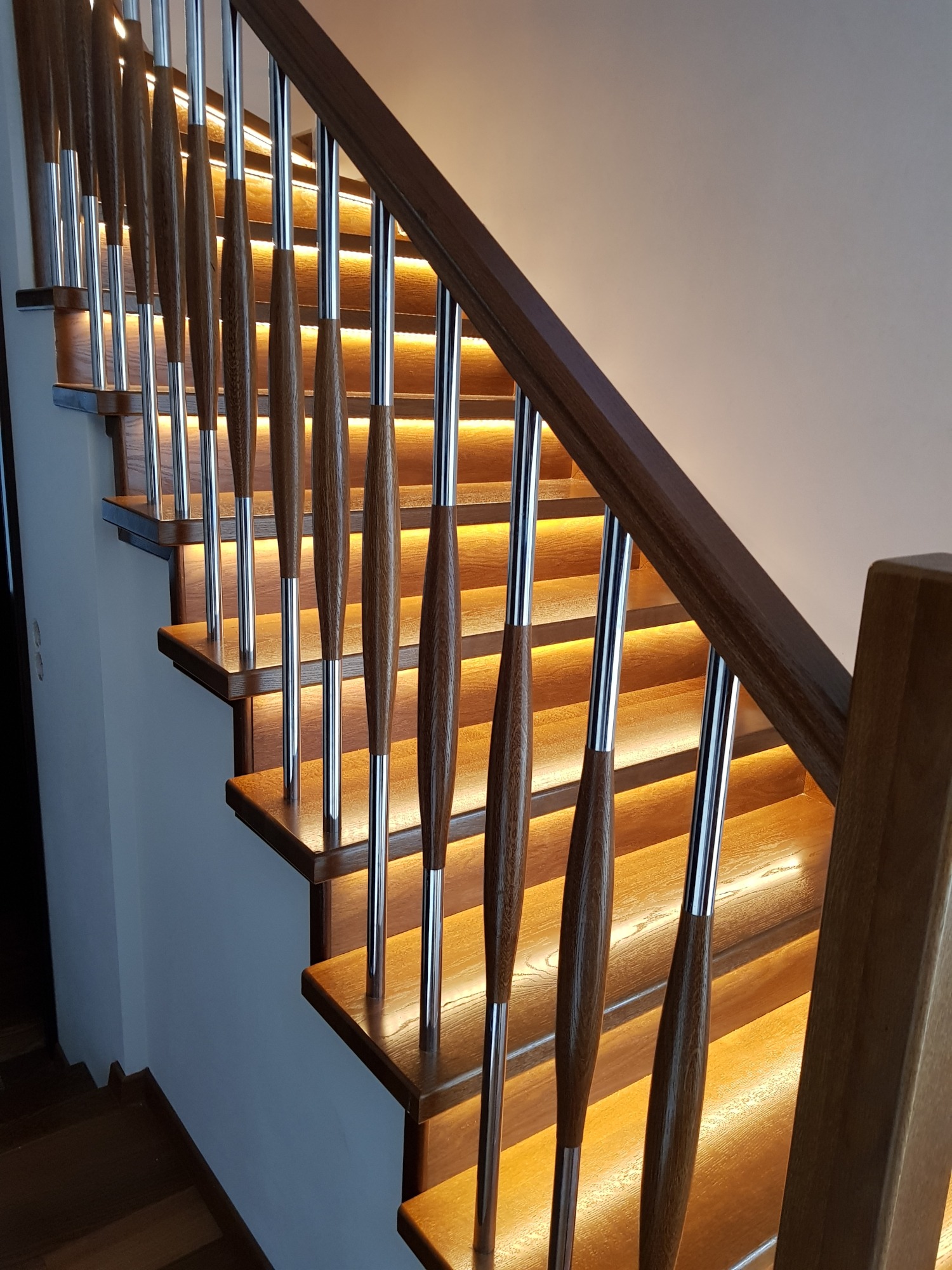 Drewniane schody z podświetleniem LED, balustrada z drewnianymi tralkami i chromowanymi elementami, widok z dołu, ciepłe oświetlenie.
