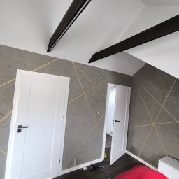 K.P. REMO - Szara ściana w pokoju na poddaszu z dekoracyjnym malowaniem złotymi liniami, białe drzwi z czarną klamką, widoczne czarne belki stropowe i fragment czerwonego fotela na podłodze.