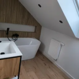 Nowoczesna łazienka na poddaszu z drewnianą ścianą, białą wanną wolnostojącą, umywalką z szafką w kolorze drewna i czarną armaturą, oświetlona światłem z okna dachowego.
