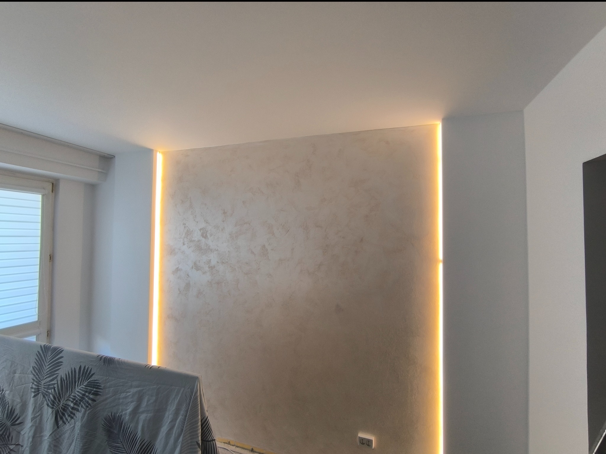 Ściana z dekoracyjną fakturą i pionowym oświetleniem LED, widoczny fragment okna z roletami i przykryty mebel z wzorem liści