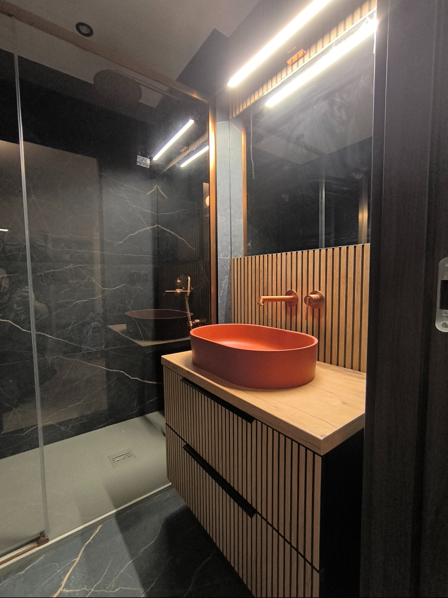 Nowoczesna łazienka z czarnymi marmurowymi płytkami, kabiną prysznicową typu walk-in i szafką z drewnianymi lamelami, na której stoi pomarańczowa umywalka nablatowa z miedzianą armaturą.