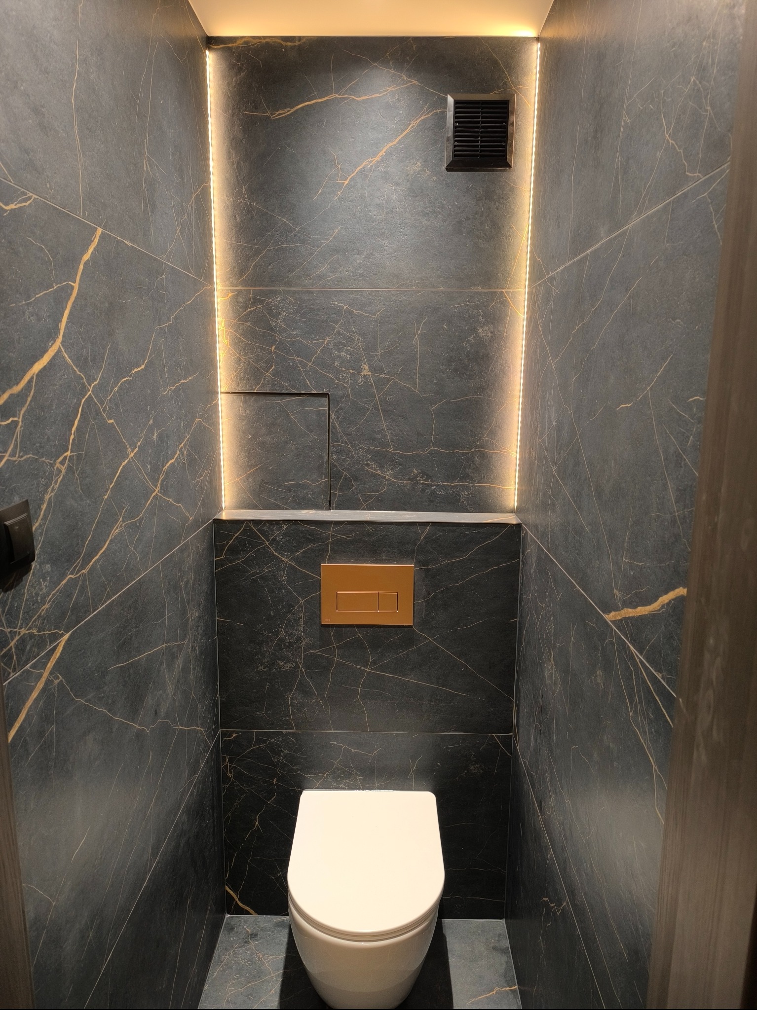 Nowoczesna toaleta w ciemnych płytkach imitujących marmur ze złotymi żyłami, podświetlona pionowymi pasami LED po bokach, widoczny panel sterujący spłuczką w kolorze miedzianym i kratka wentylacyjna.