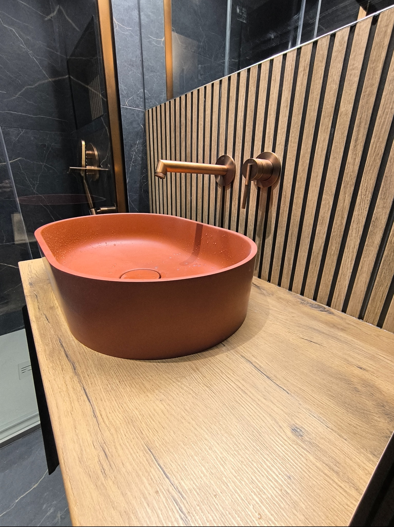 Nowoczesna łazienka z umywalką w kolorze terakoty, armaturą w odcieniu miedzi i ścianą z drewnianych paneli o pionowym układzie, uzupełniona czarnymi płytkami imitującymi marmur w kabinie prysznicowej
