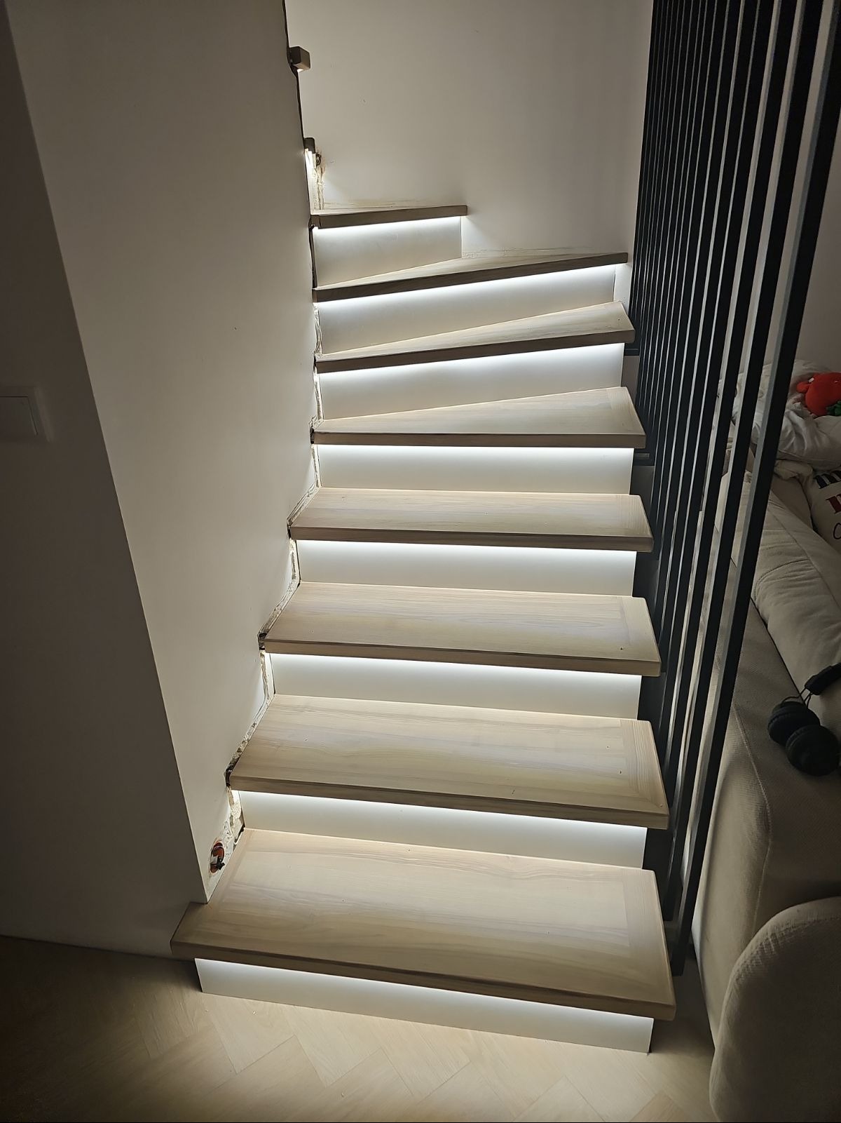 Drewniane schody spiralne z podświetleniem LED, widoczne niedokończone wykończenie przy ścianie, czarna barierka, fragment sofy w tle.