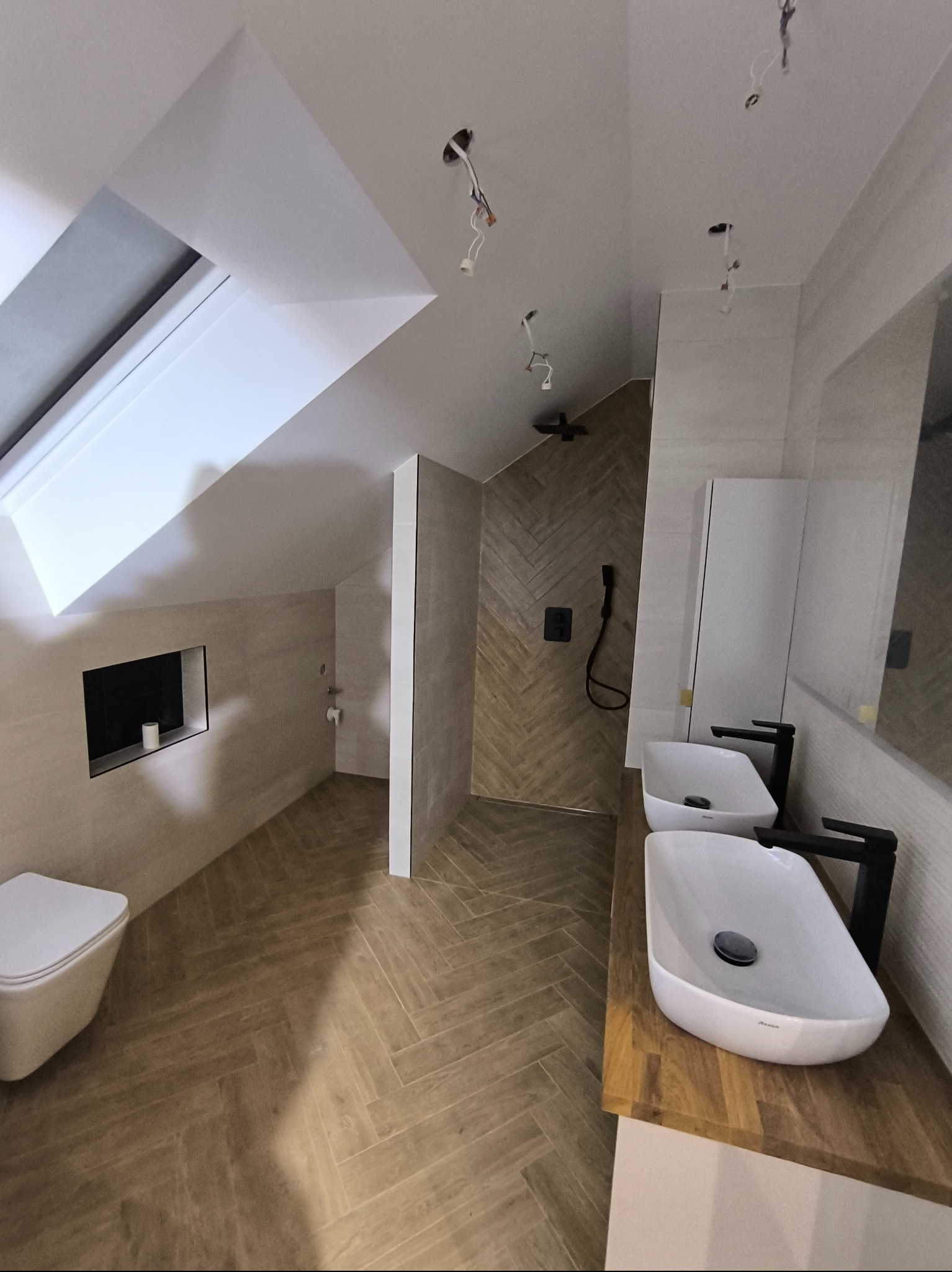 Nowoczesna łazienka na poddaszu z drewnopodobną podłogą ułożoną w jodełkę, dwie białe umywalki nablatowe z czarnymi bateriami, toaleta pod skosem i kabina prysznicowa z deszczownicą.