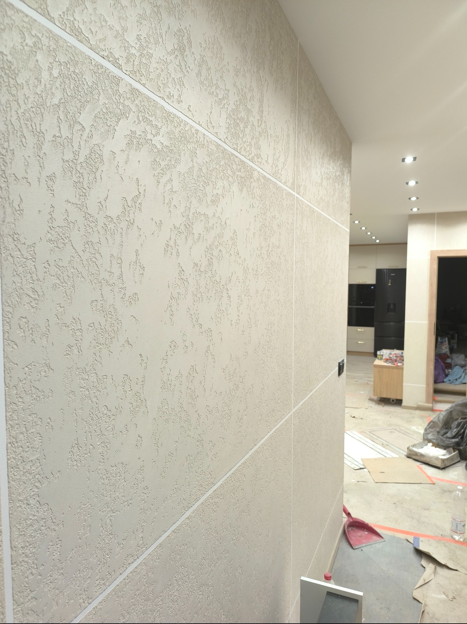 Beżowa ściana z dekoracyjnym tynkiem strukturalnym, podzielona białymi listwami, widok na wnętrze w trakcie prac wykończeniowych z widocznymi narzędziami i zabezpieczeniami podłogi.