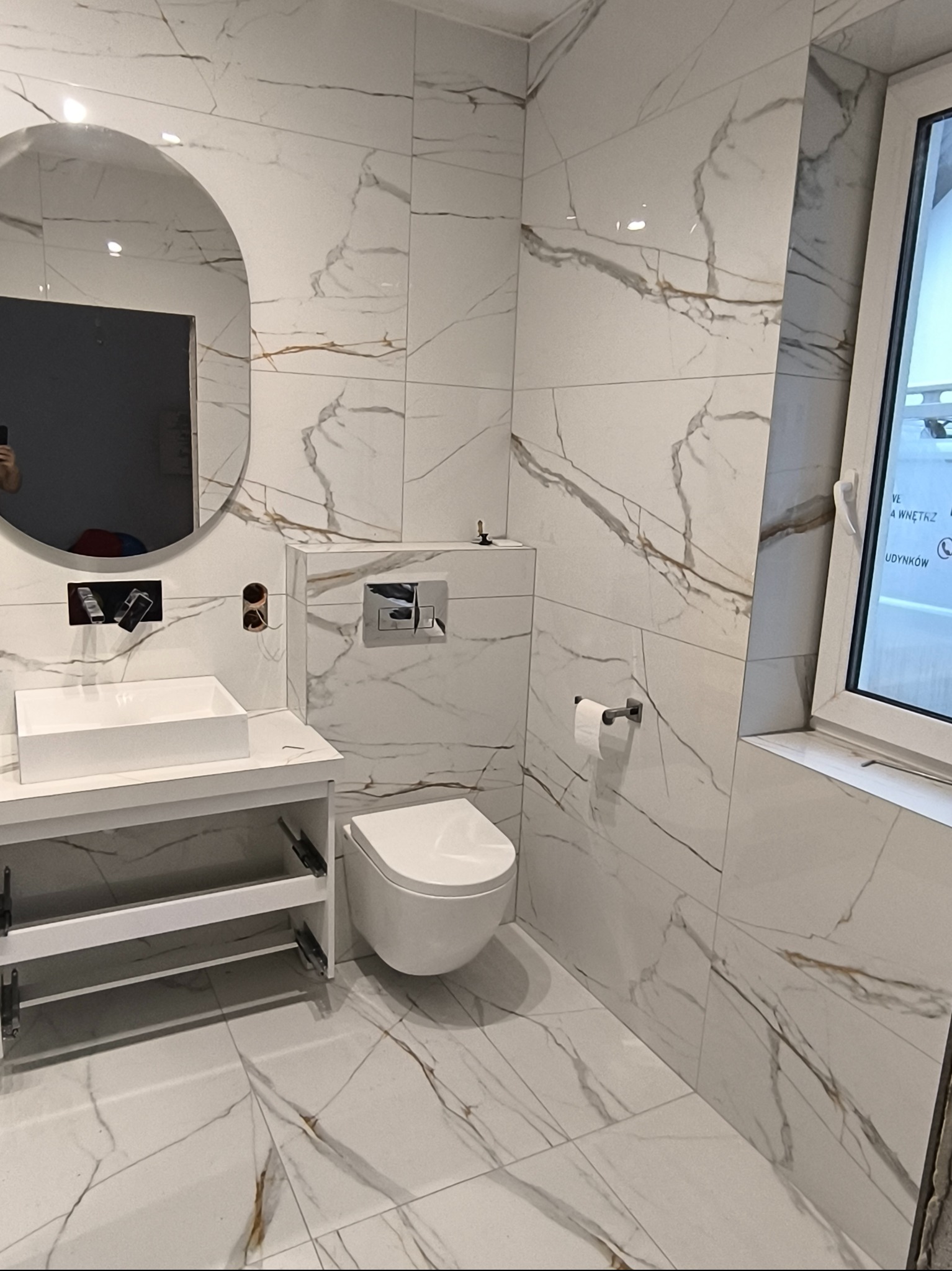 Nowoczesna łazienka z białymi płytkami imitującymi marmur, owalne lustro, wisząca toaleta i biała umywalka na szafce z otwartymi półkami, widoczne elementy instalacji przed montażem baterii.