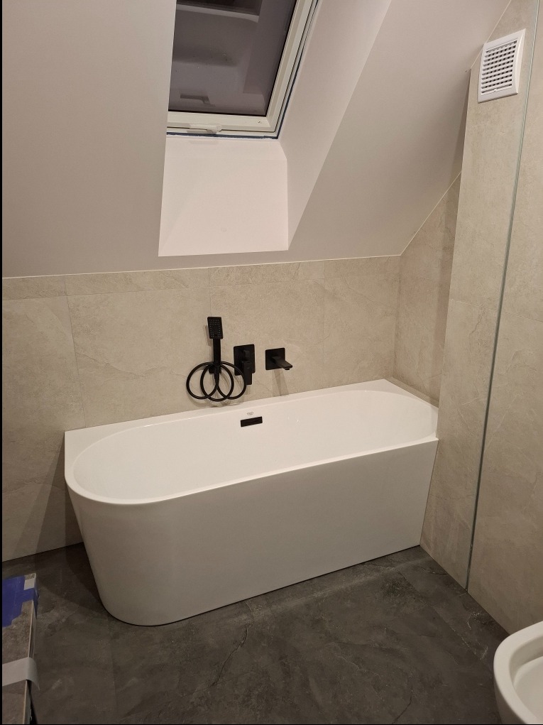 Nowoczesna, biała wanna wolnostojąca z czarną armaturą na tle beżowych płytek w łazience na poddaszu ze skośnym oknem dachowym.