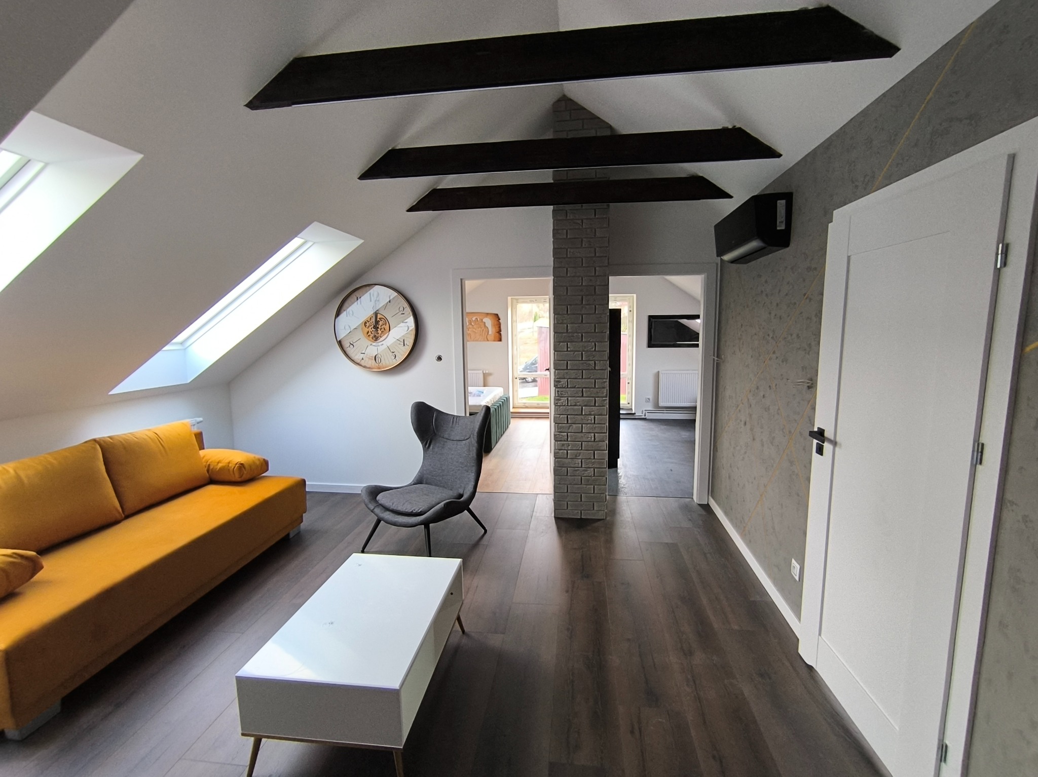 Jasne, przestronne wnętrze poddasza z żółtą sofą, szarym fotelem, białym stolikiem kawowym i drewnianą podłogą. Widoczne okna dachowe, ceglana kolumna i czarne belki dekoracyjne na suficie.
