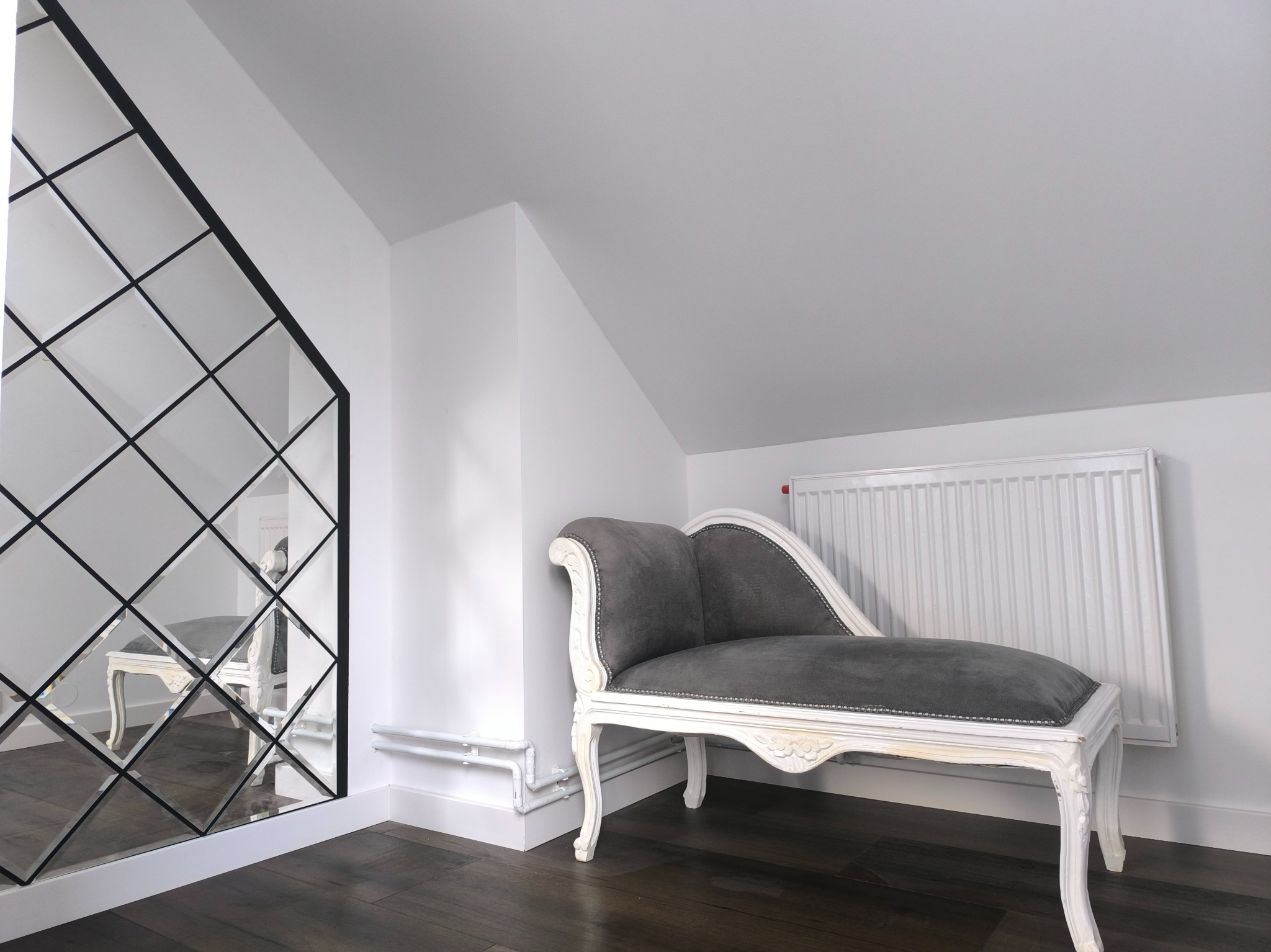 Białe wnętrze poddasza z dekoracyjnym lustrem w czarnej ramie, szarą leżanką w stylu Ludwika i grzejnikiem.