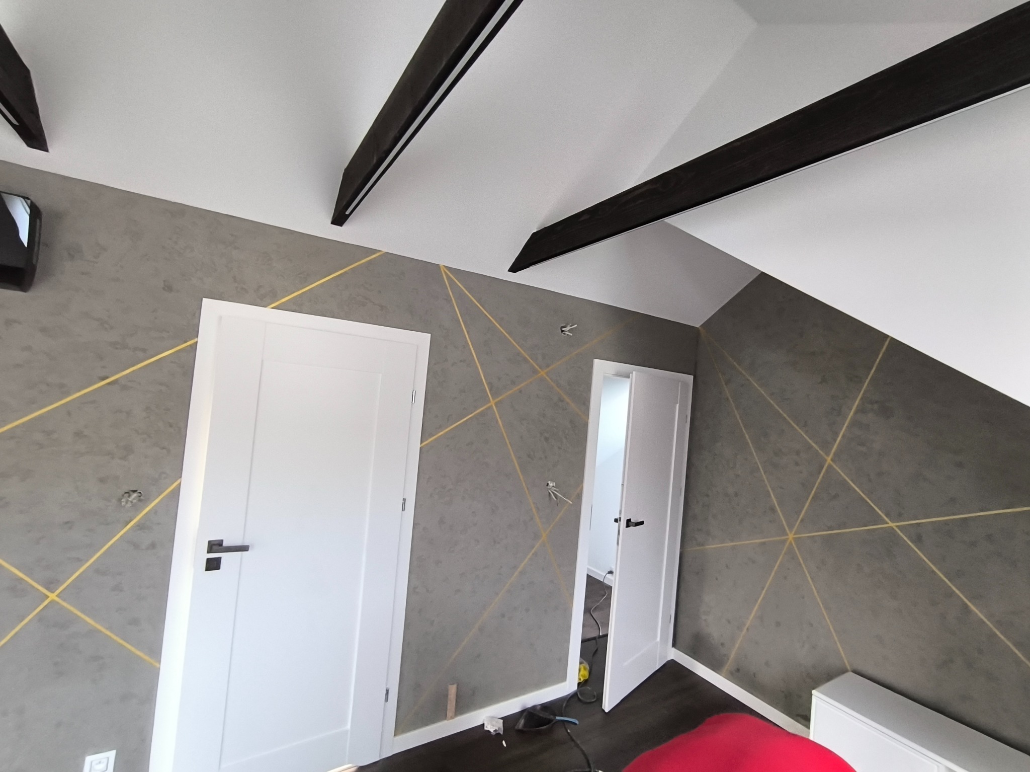 Szara ściana w pokoju na poddaszu z dekoracyjnym malowaniem złotymi liniami, białe drzwi z czarną klamką, widoczne czarne belki stropowe i fragment czerwonego fotela na podłodze.