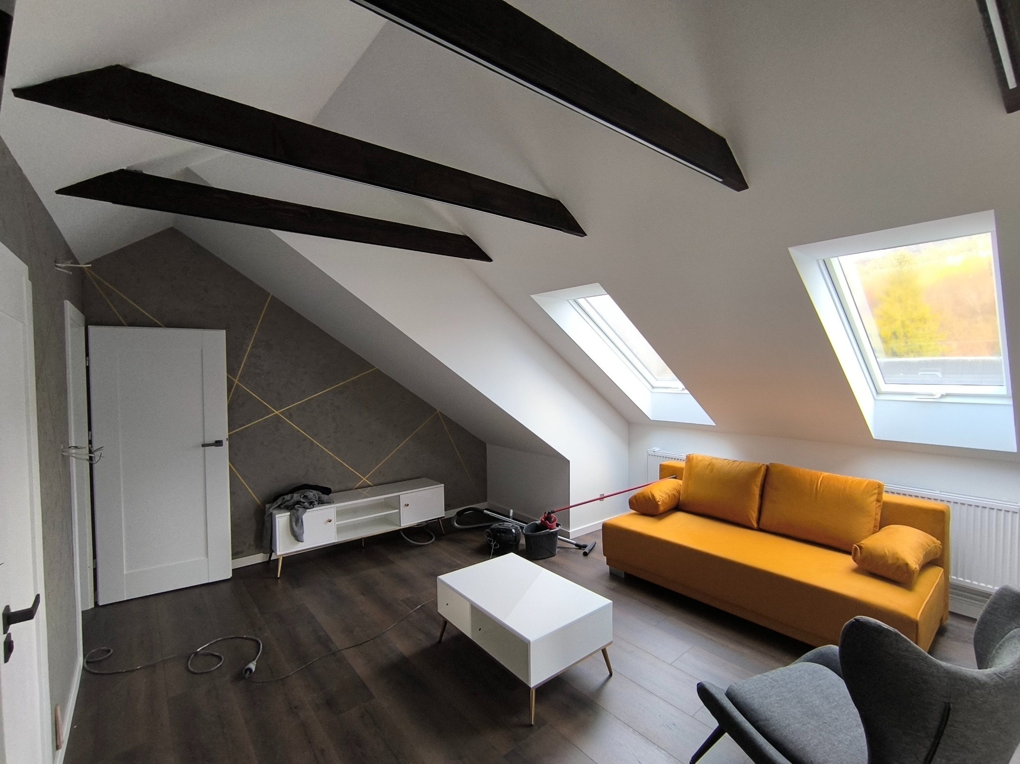 Pokój na poddaszu z skośnym sufitem, drewnianymi belkami, żółtą sofą, białym stolikiem kawowym i szarą tapetą z geometrycznym wzorem, częściowo umeblowany, w trakcie wykańczania.