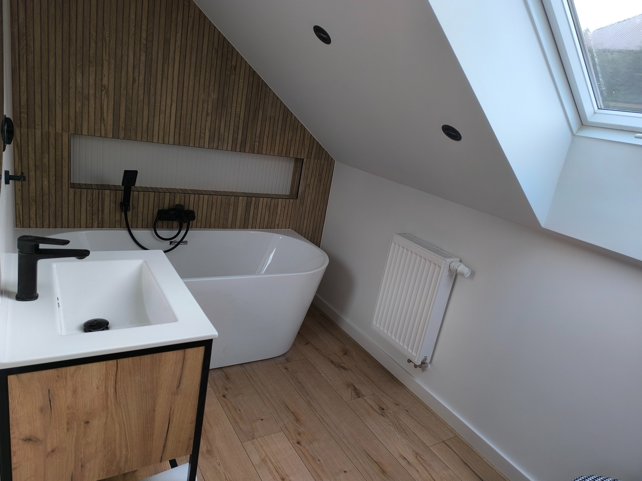 Nowoczesna łazienka na poddaszu z drewnianą ścianą, białą wanną wolnostojącą, umywalką z szafką w kolorze drewna i czarną armaturą, oświetlona światłem z okna dachowego.