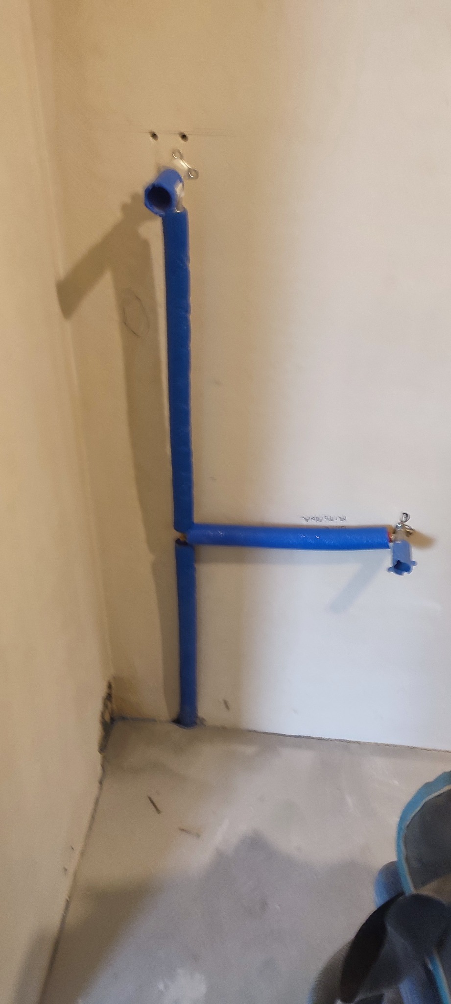 Niebieskie rury wodne połączone w kształt litery L, zamontowane na ścianie w surowym stanie, widoczne mocowania i fragment podłogi, ujęcie z dołu.