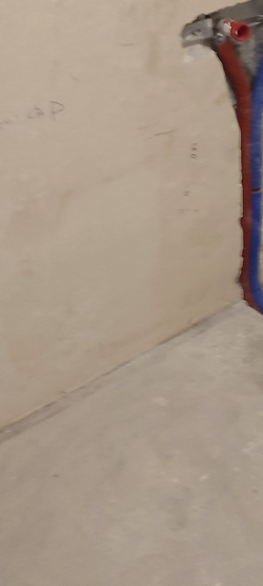 Fragment ściany z wystającymi rurami instalacji wodnej w kolorze czerwonym i niebieskim, zamontowanymi w metalowym uchwycie, oraz surowym wykończeniem