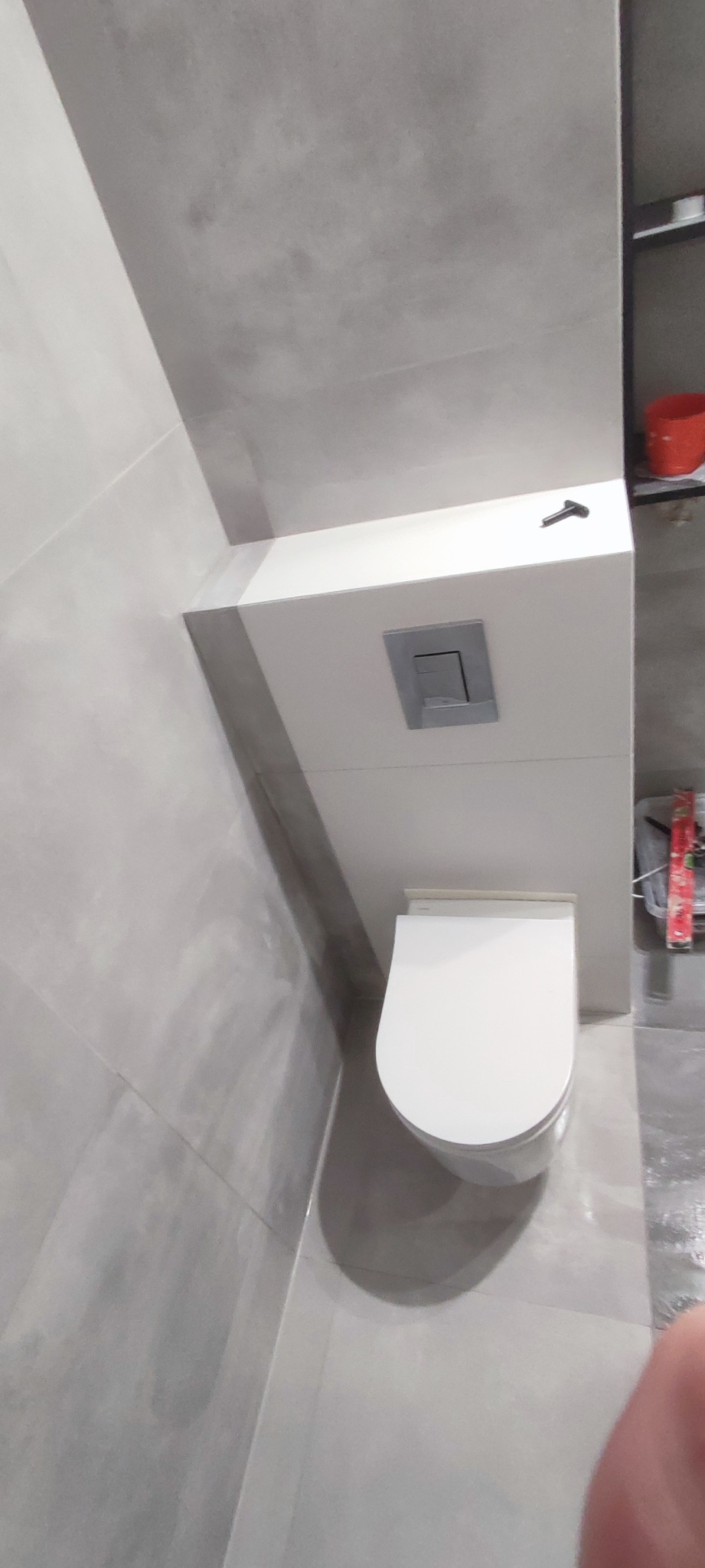 Nowoczesna, podwieszana toaleta z szarymi płytkami w tle, widoczny chromowany przycisk spłukiwania i poziomica w tle