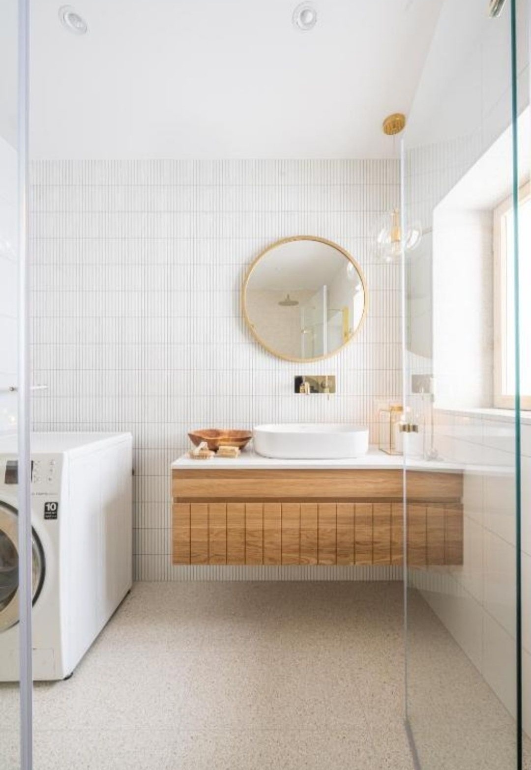 Nowoczesna łazienka z białą armaturą, umywalką nablatową na drewnianej szafce, okrągłym lustrem w złotej ramie i pralką w tle