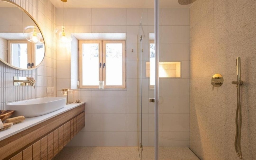 Nowoczesna łazienka z drewnianą szafką pod umywalką, okrągłym lustrem w złotej ramie, szklanym prysznicem i złotą armaturą, oświetlona wiszącymi lampami z przezroczystymi kloszami.