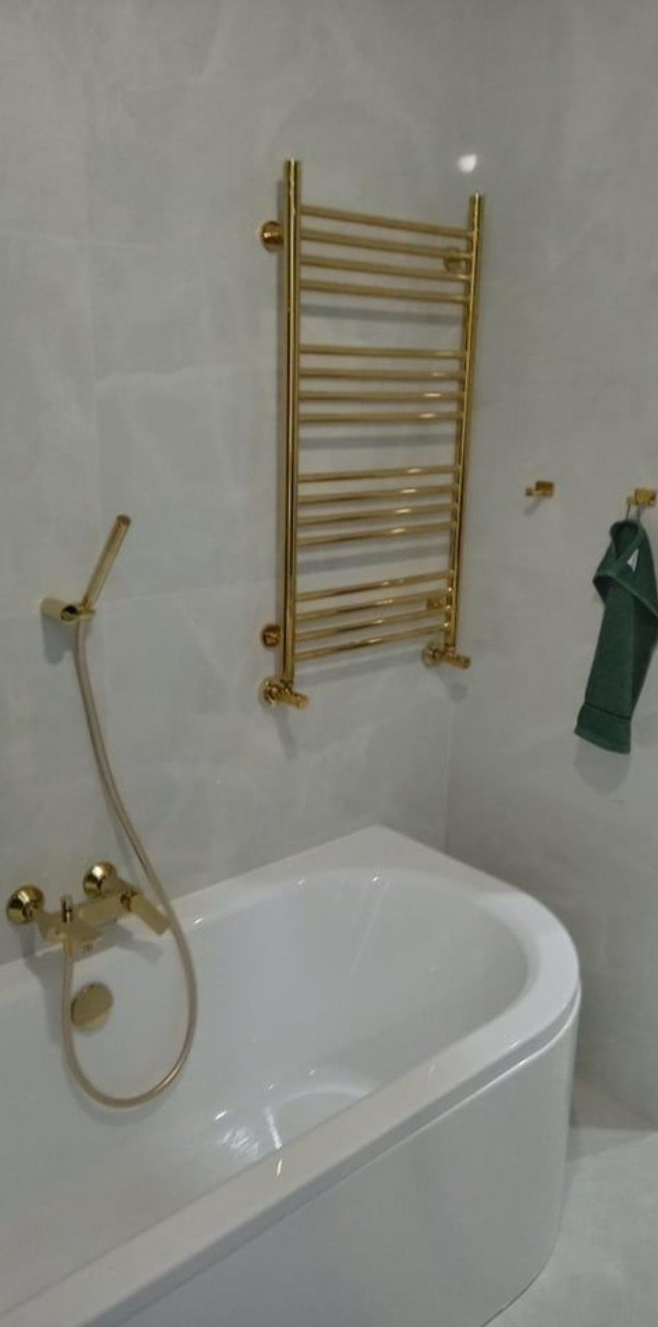 Biała wanna narożna z baterią prysznicową w kolorze złotym i grzejnik drabinkowy łazienkowy w tym samym kolorze, ręcznik w odcieniach zieleni.