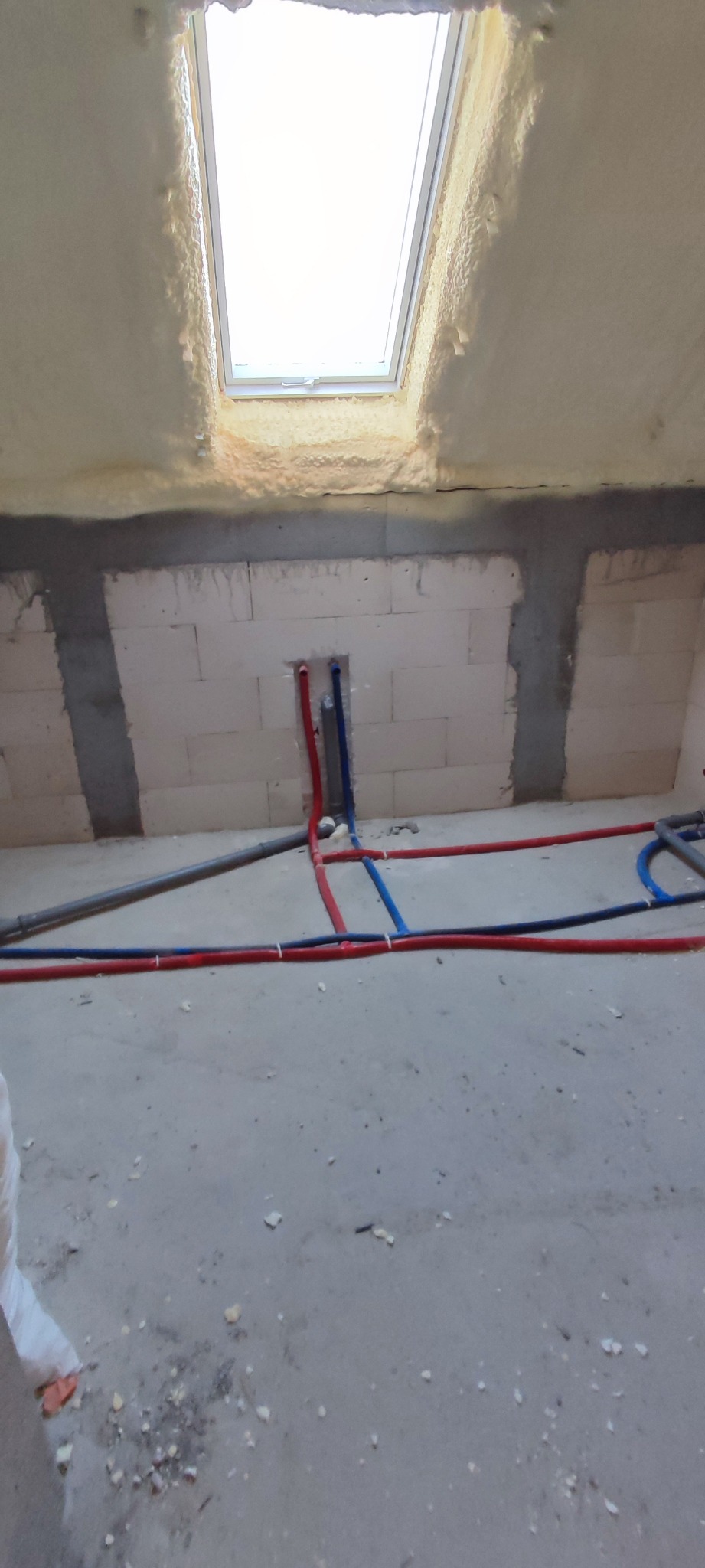 Surowy stan poddasza z oknem dachowym, izolacją piankową i częściowo rozprowadzonymi rurami instalacji wodnej w kolorach czerwonym i niebieskim.