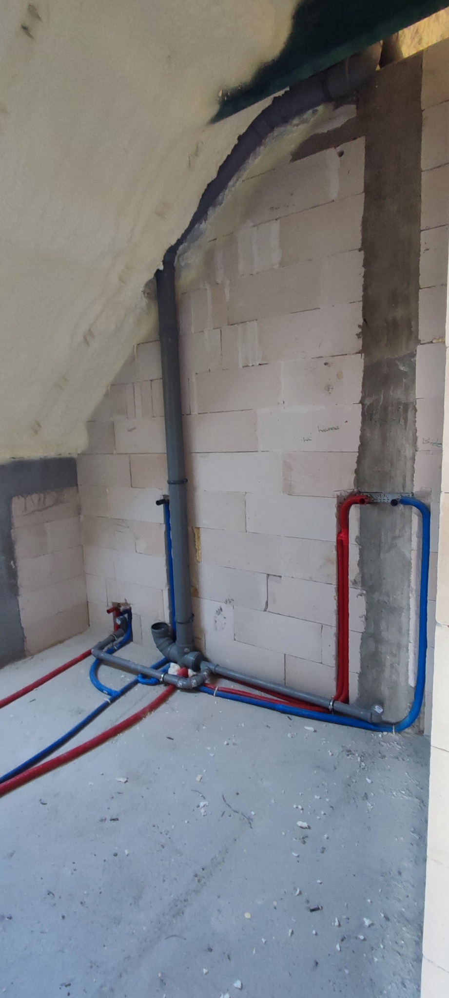 Surowy stan wykończenia poddasza z widocznymi rurami instalacji wodnej i kanalizacyjnej w kolorze szarym, czerwonym i niebieskim, prowadzącymi wzdłuż ścian z betonu komórkowego.