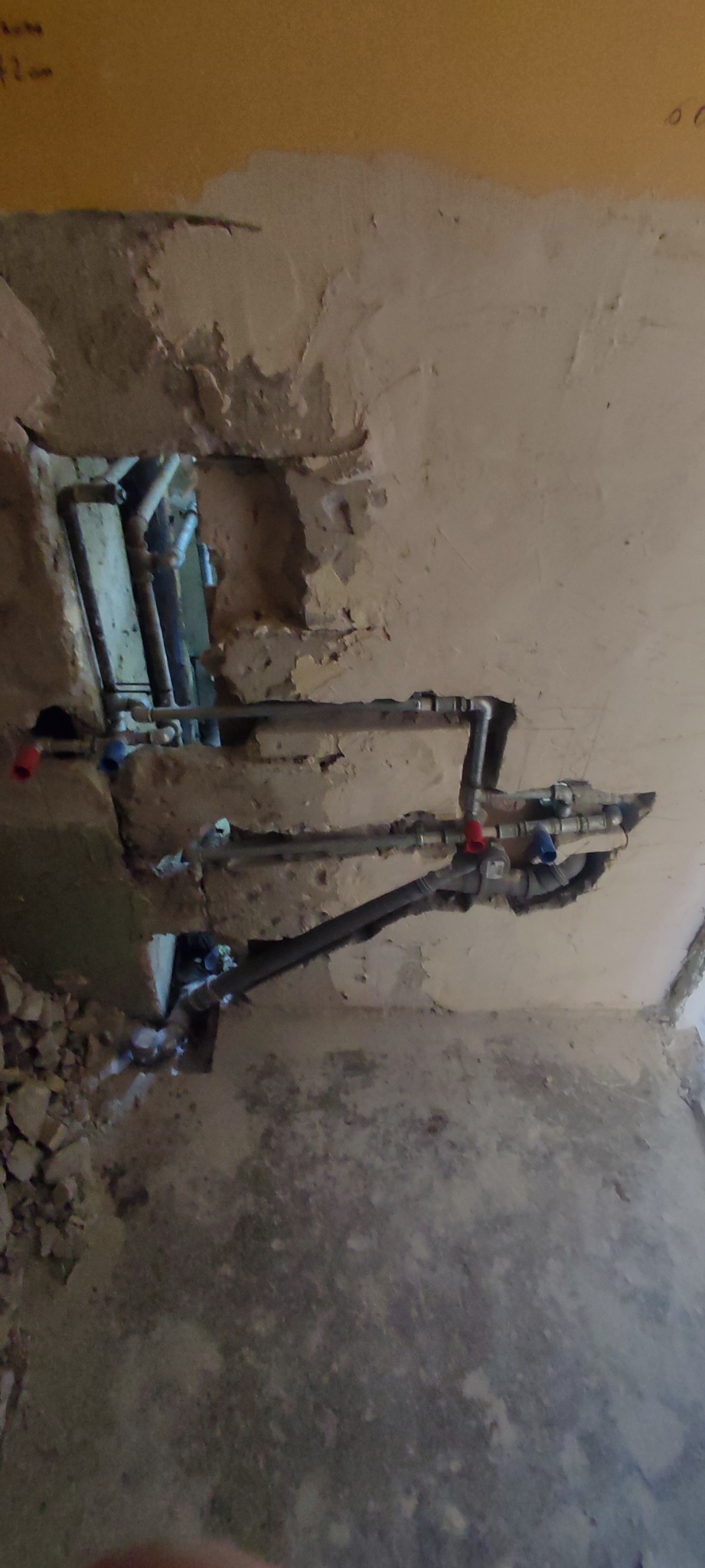 Fragment ściany z odsłoniętymi rurami instalacji wodnej z zaworami, widoczne ślady po kuciu w tynku, w tle inne rury