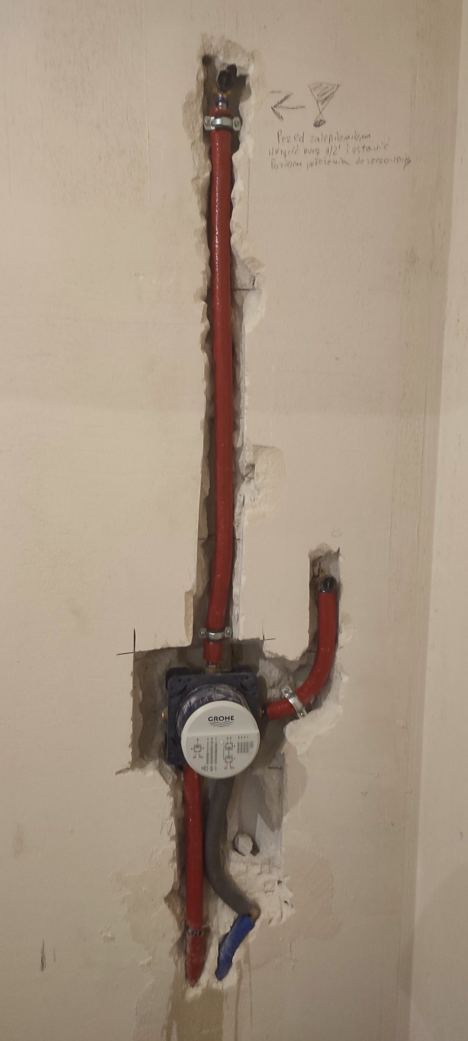 Czerwone rury wodne zamontowane w ścianie z licznikiem Grohe, widoczne ślady po kuciu w ścianie, notatka ołówkiem na ścianie z instrukcjami.
