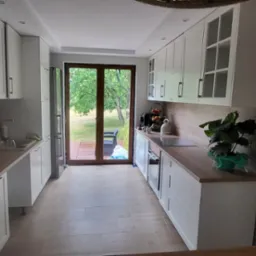 Jasna kuchnia z białymi szafkami, drewnianymi blatami i widokiem na ogród przez przeszklone drzwi balkonowe.