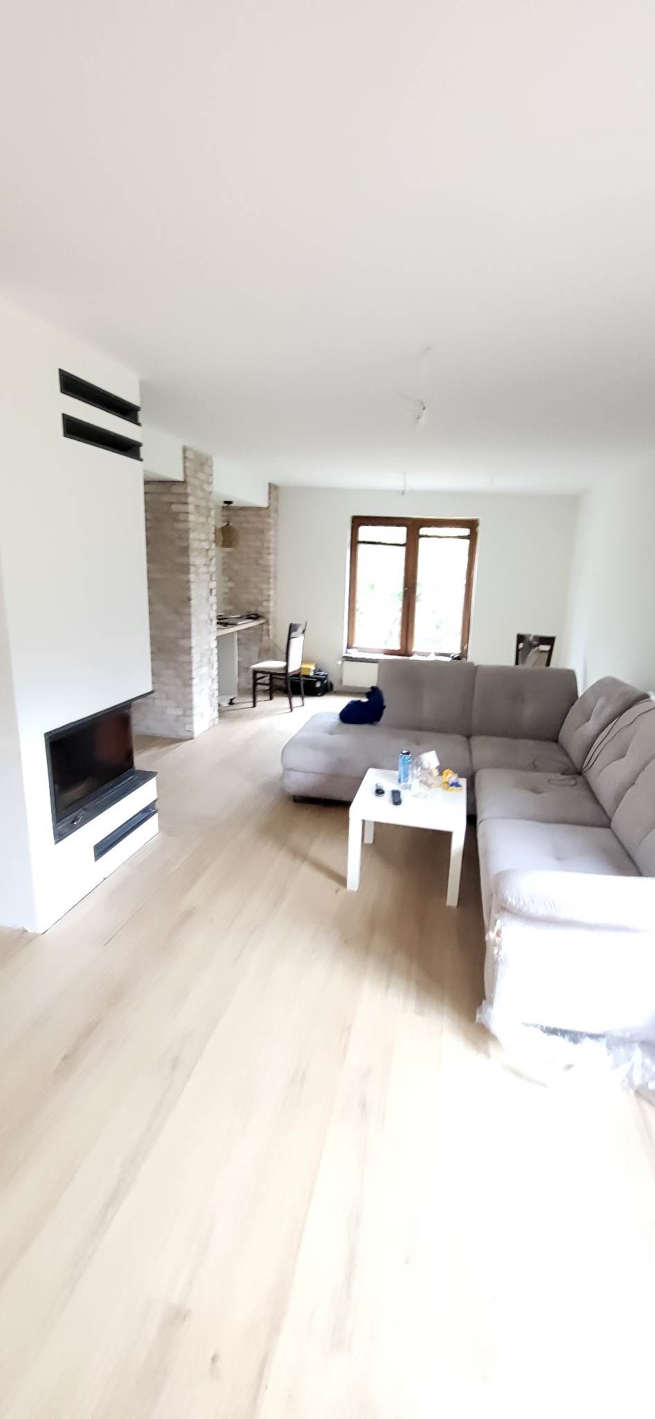 Jasne panele podłogowe w salonie z szarą sofą narożną, białym stolikiem kawowym i kominkiem z czarnym wkładem, w tle ściana z dekoracyjnej cegły i częściowo widoczna kuchnia.