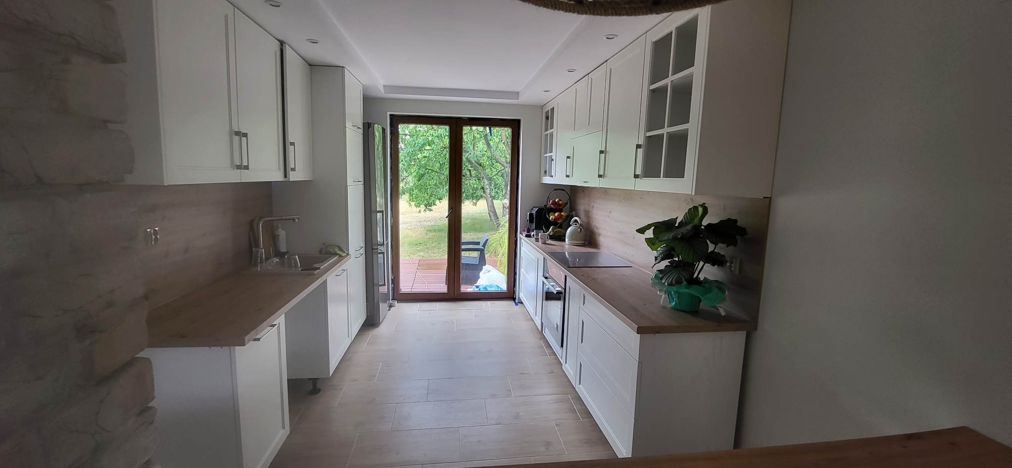 Jasna kuchnia z białymi szafkami, drewnianymi blatami i widokiem na ogród przez przeszklone drzwi balkonowe.