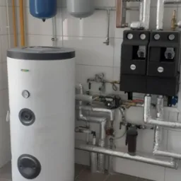 Biały zasobnik ciepłej wody użytkowej z podłączonymi rurami, zaworami i pompami obiegowymi w kotłowni z szarą podłogą z płytek.