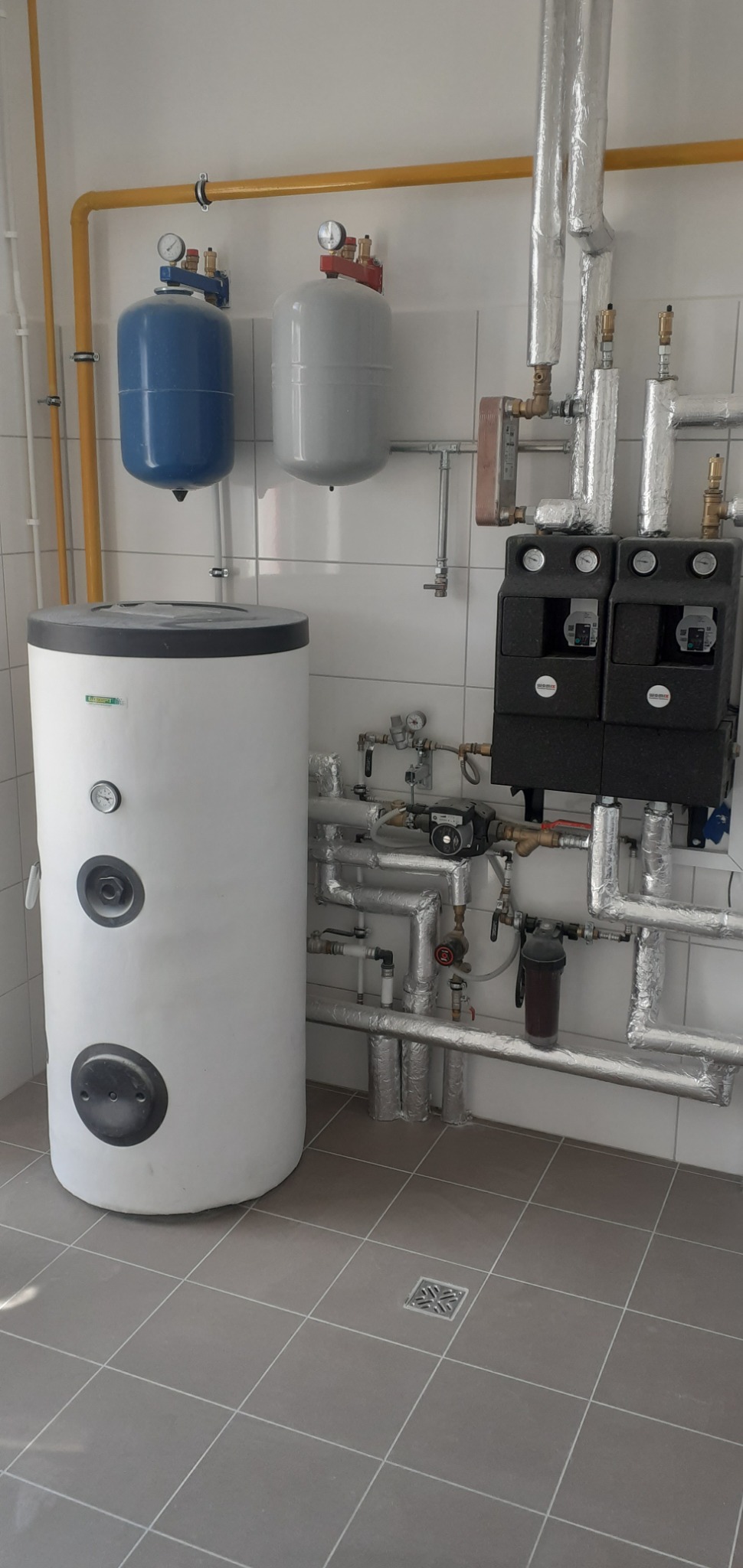 Biały zasobnik ciepłej wody użytkowej z podłączonymi rurami, zaworami i pompami obiegowymi w kotłowni z szarą podłogą z płytek.