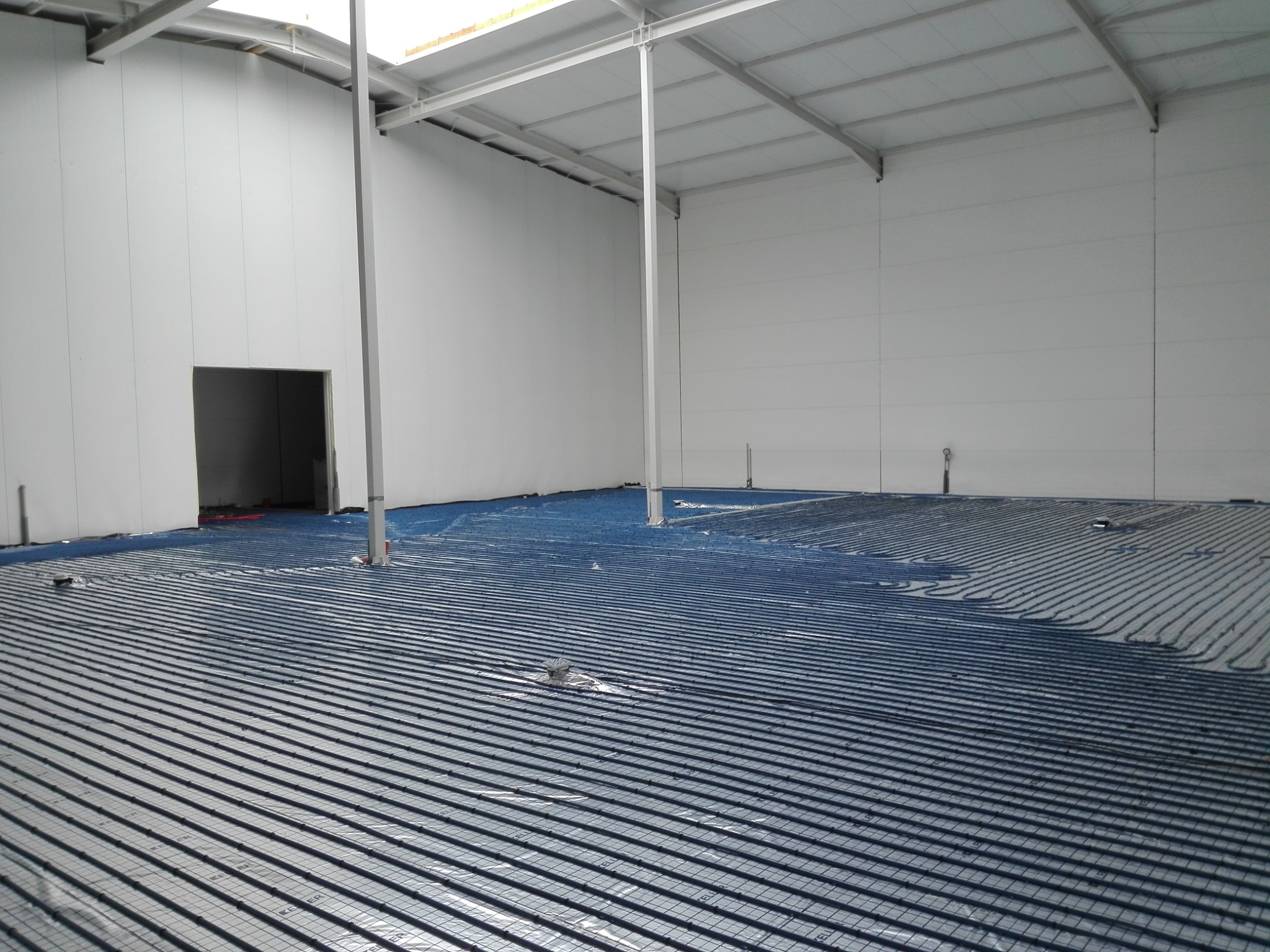 Rozległa hala z widoczną instalacją ogrzewania podłogowego, rury ułożone w regularnych odstępach na folii odblaskowej, przygotowane do zalania wylewką.