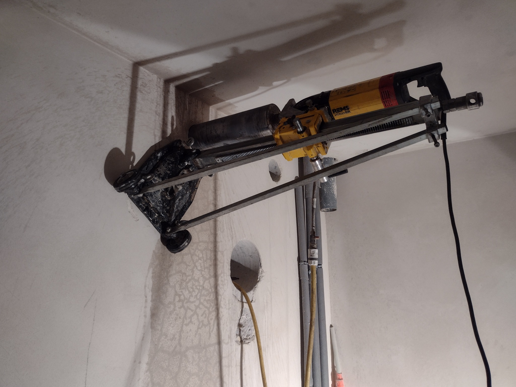 Wiertnica diamentowa REMS RIDGID SR w trakcie wiercenia otworu w ścianie, widoczne instalacje rurowe i przewód zasilający wiertnicę.