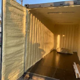 Wnętrze kontenera po natrysku pianką poliuretanową, widoczne r&oacute;wnomierne pokrycie ścian, sufitu i otwartych drzwi, podłoga wyłożona panelami, w tle budynki mieszkalne.