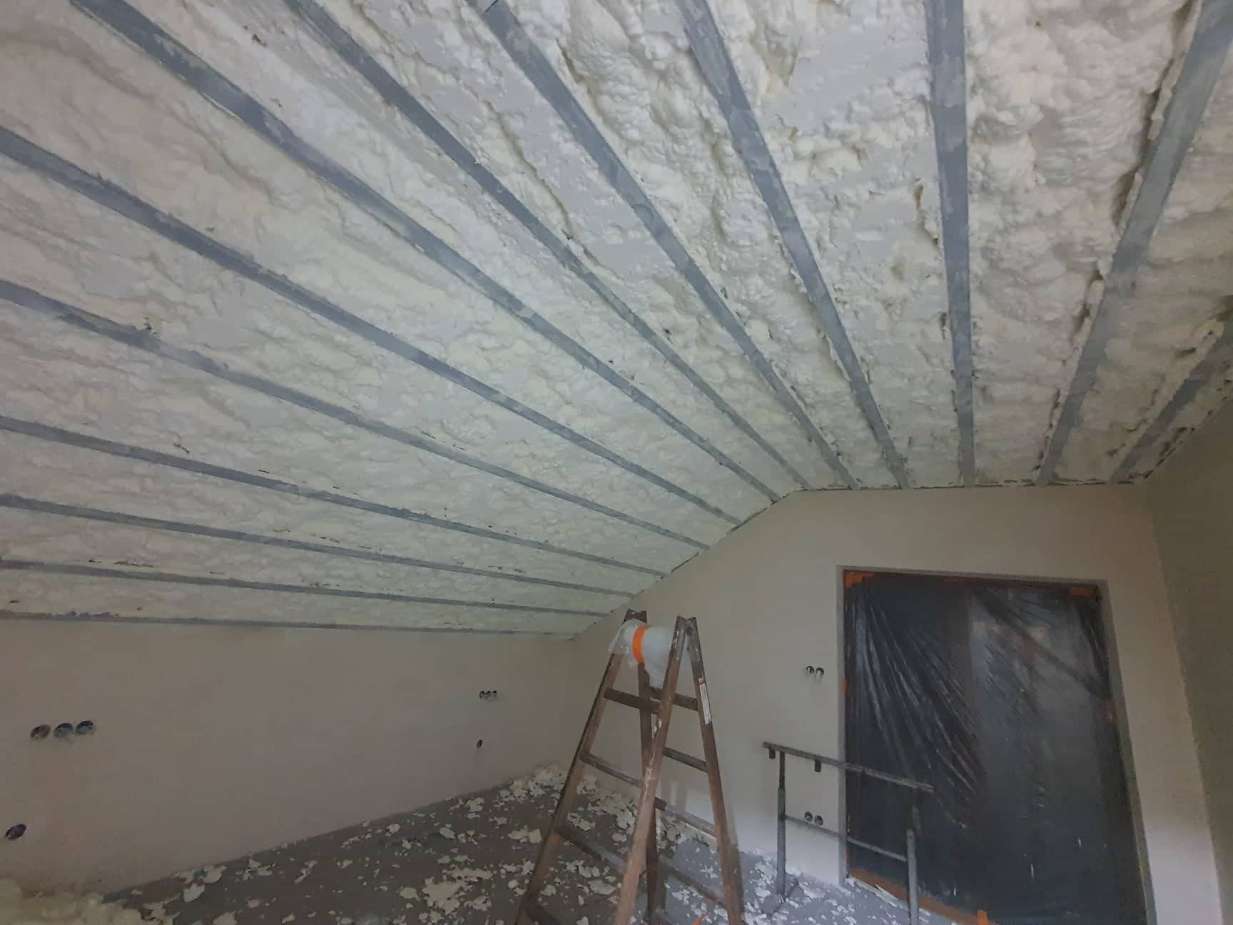 Poddasze po ociepleniu pianką PUR, widoczne metalowe belki konstrukcyjne pokryte pianą, drabina oparta o ścianę, zabezpieczone folią drzwi.