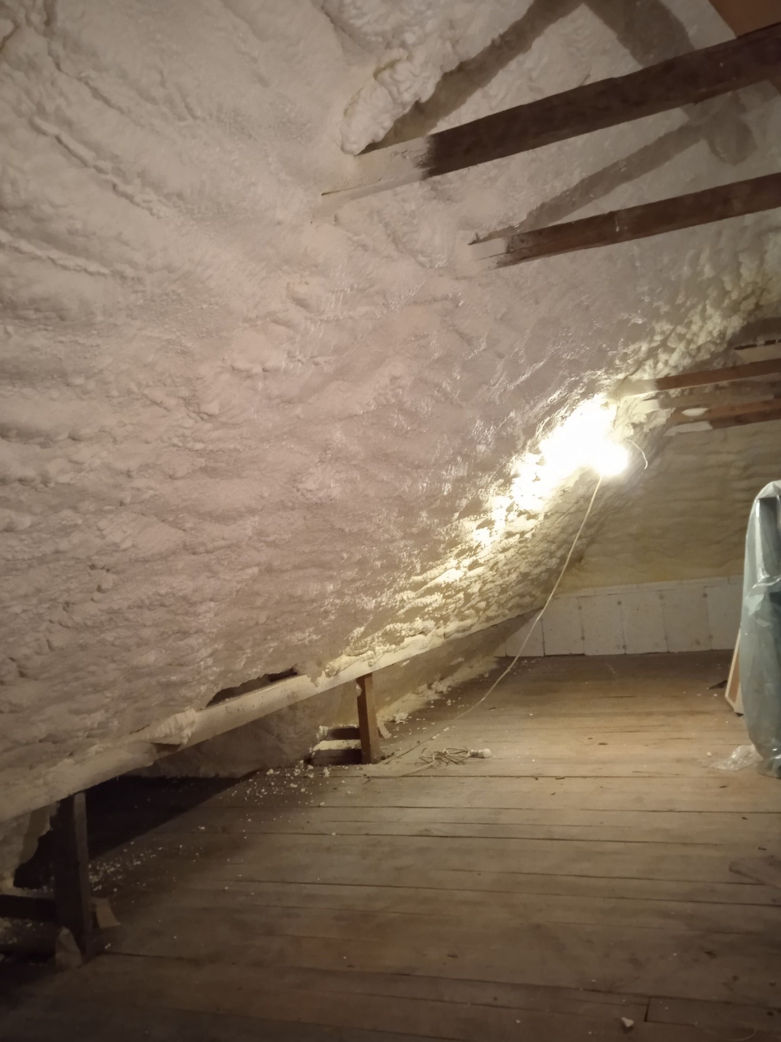 Poddasze w Ostrowi Mazowieckiej po ociepleniu pianką poliuretanową, widoczne drewniane belki stropowe częściowo pokryte pianą, oraz oświetlenie robocze.